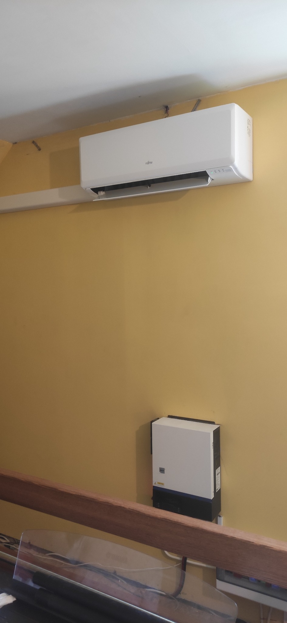 Biały klimatyzator marki Fujitsu zamontowany na żółtej ścianie, poniżej widoczny moduł inwertera, a w dolnej części kadru fragment balustrady.