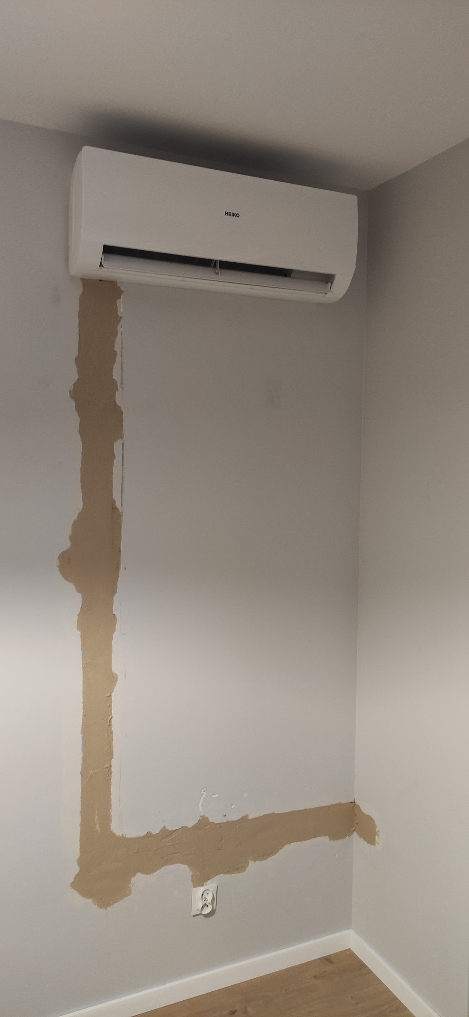 Biały klimatyzator marki Heiko zamontowany na ścianie z widocznymi śladami po montażu i naprawie, z fragmentami ściany pokrytymi świeżą szpachlą w kształcie litery L.