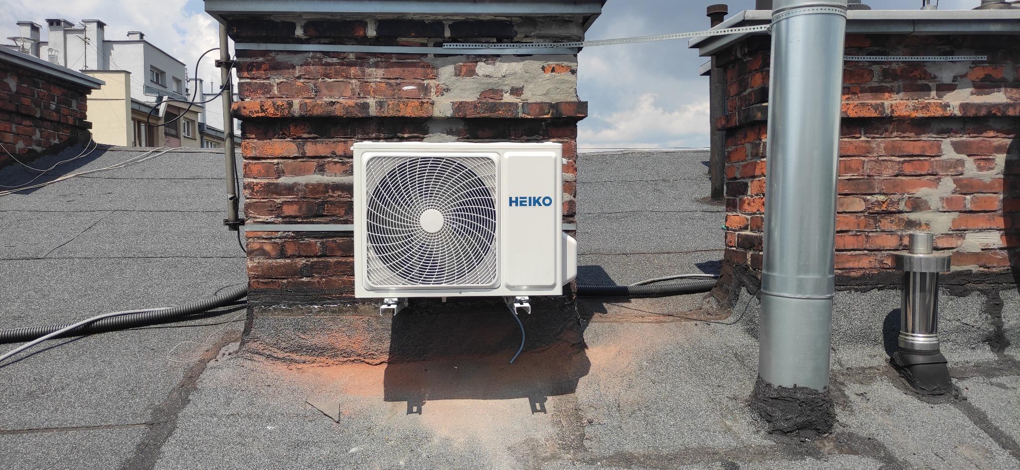 Zewnętrzna jednostka klimatyzacji marki HEIKO zamontowana na dachu budynku, widoczna instalacja i przewody, w tle kominy i budynki mieszkalne.