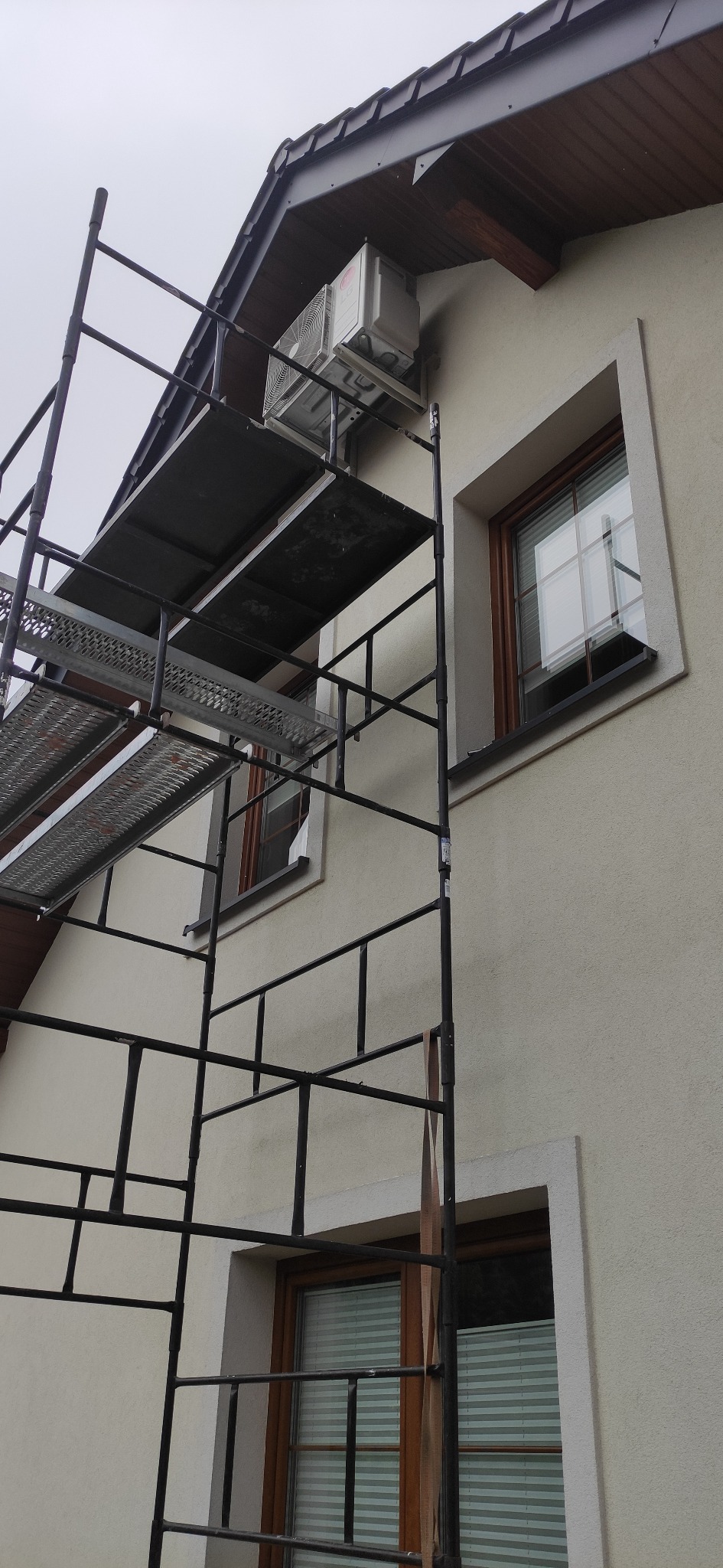 Zewnętrzna jednostka klimatyzacji zamontowana na ścianie domu, widoczna z poziomu rusztowania. Ściana w jasnym odcieniu z dwoma oknami z brązowymi ramami.