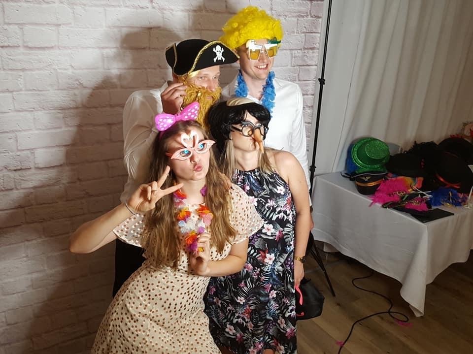 Grupa czterech osób pozuje do zdjęcia w fotobudce, ubrani w peruki, okulary i inne rekwizyty na tle ściany z cegieł.