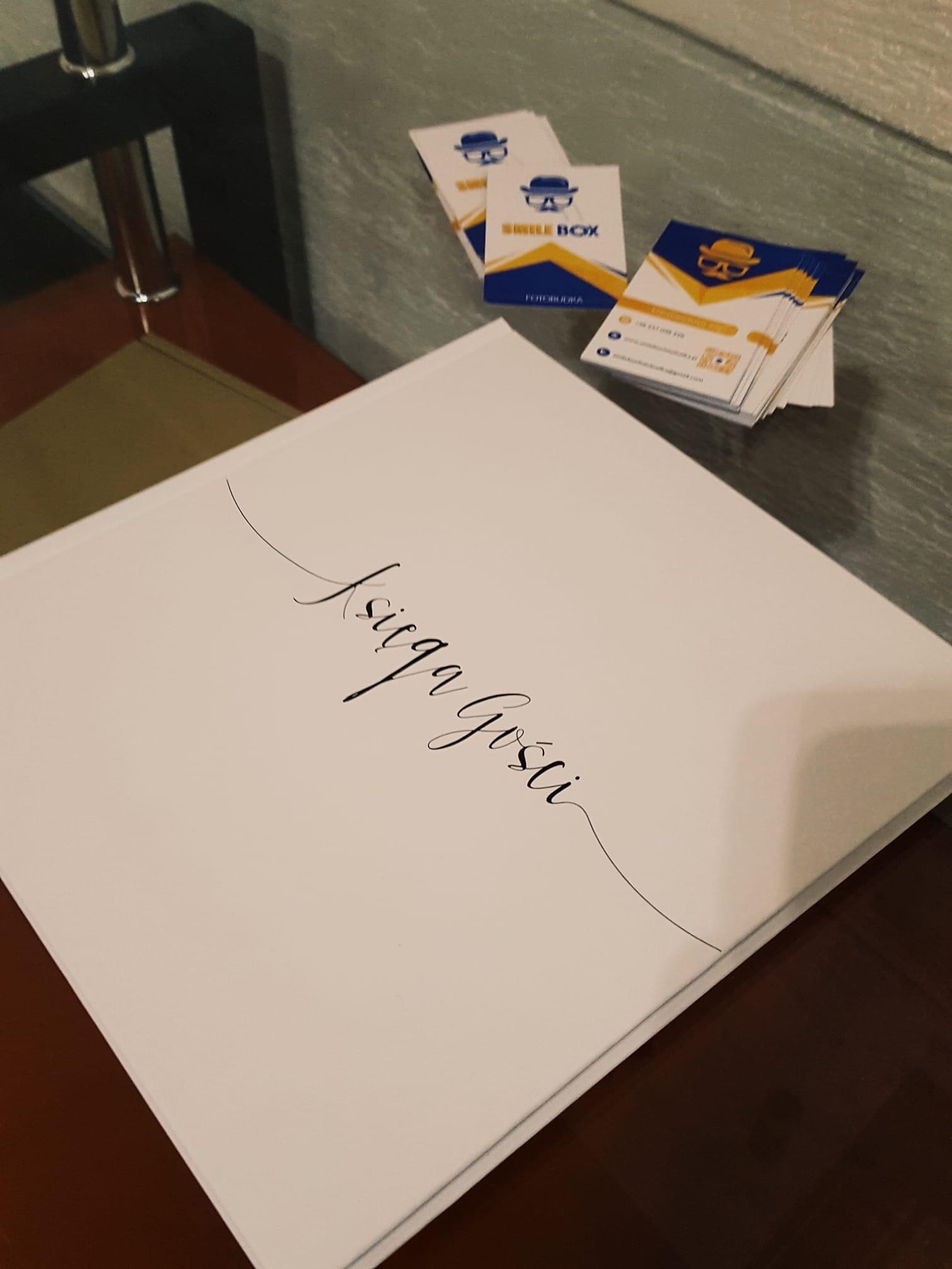 Księga gości z napisem wykonanym ozdobną czcionką, obok wizytówki firmy oferującej fotobudki.