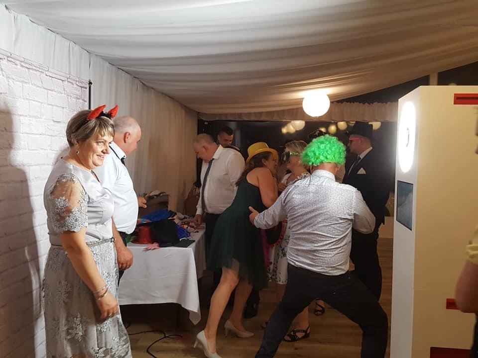 Grupa osób w przebraniach pozuje do zdjęcia w fotobudce na tle białych zasłon i ceglanej ściany.