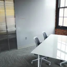 Pusta sala konferencyjna z białym stołem, szarymi krzesłami na kółkach i wzorzystą wykładziną; ściana z napisem 'Schwary' i odsłoniętą cegłą przy oknie.