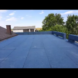 Niebieski dach pokryty płytami z widocznym kominem budynku obok i budynkiem gospodarczym w tle.