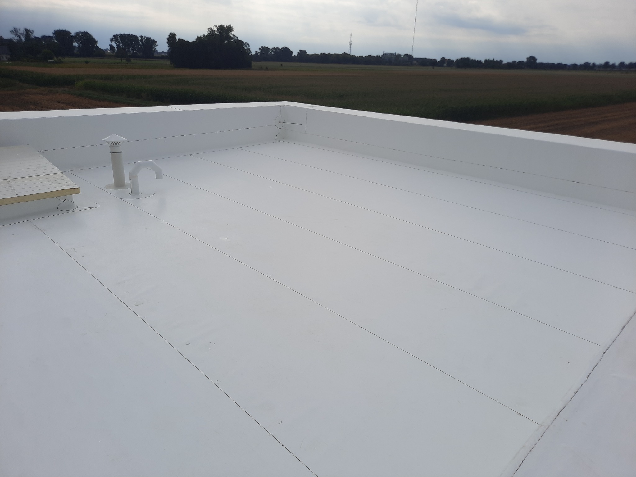 Biały, płaski dach z widocznymi łączeniami papy, kominkiem wentylacyjnym i wyjściem na dach, na tle pola uprawnego pod pochmurnym niebem.