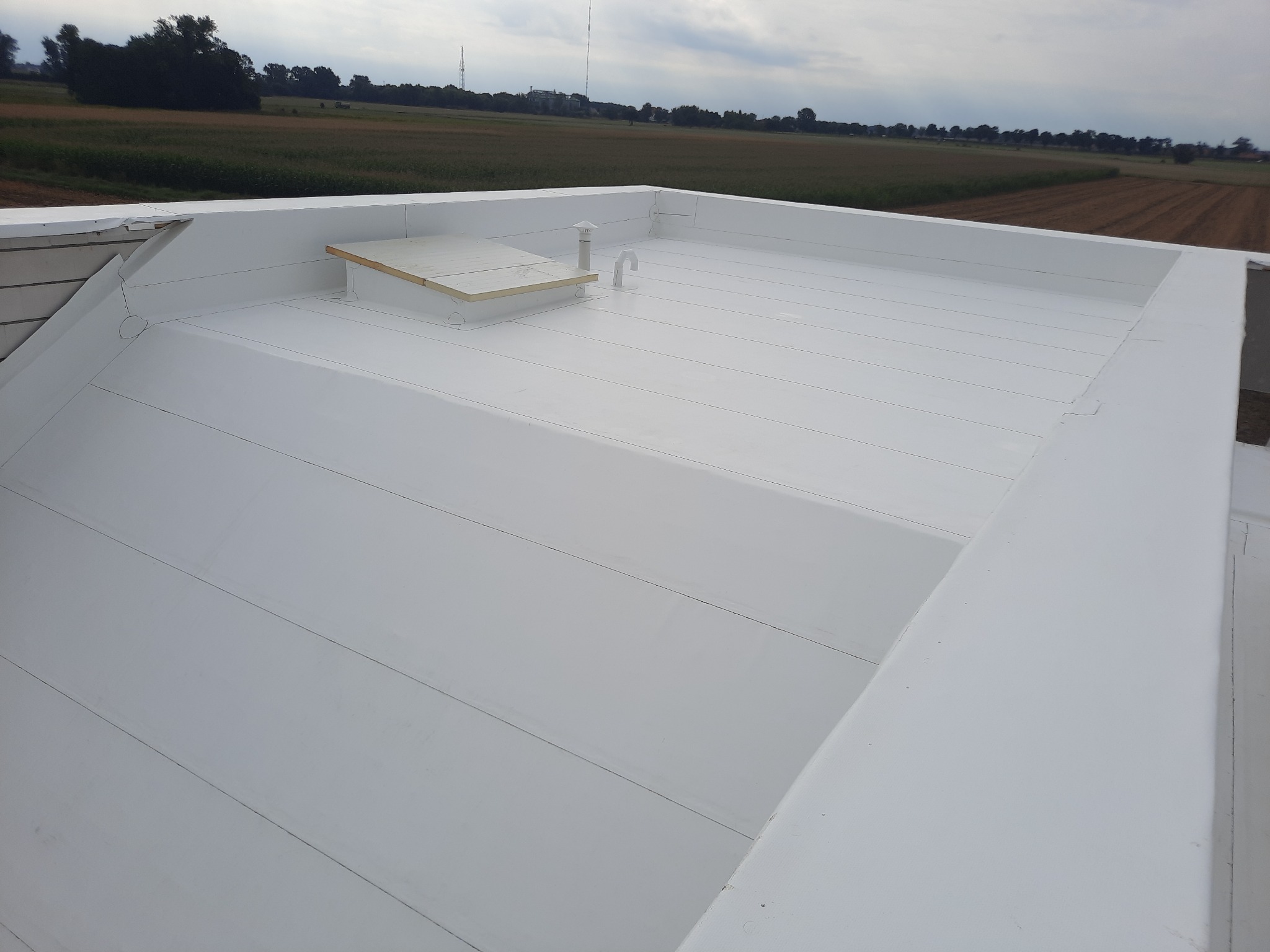 Płaski, biały dach z widocznym świetlikiem, kominkiem wentylacyjnym i tłem z polami uprawnymi pod pochmurnym niebem.