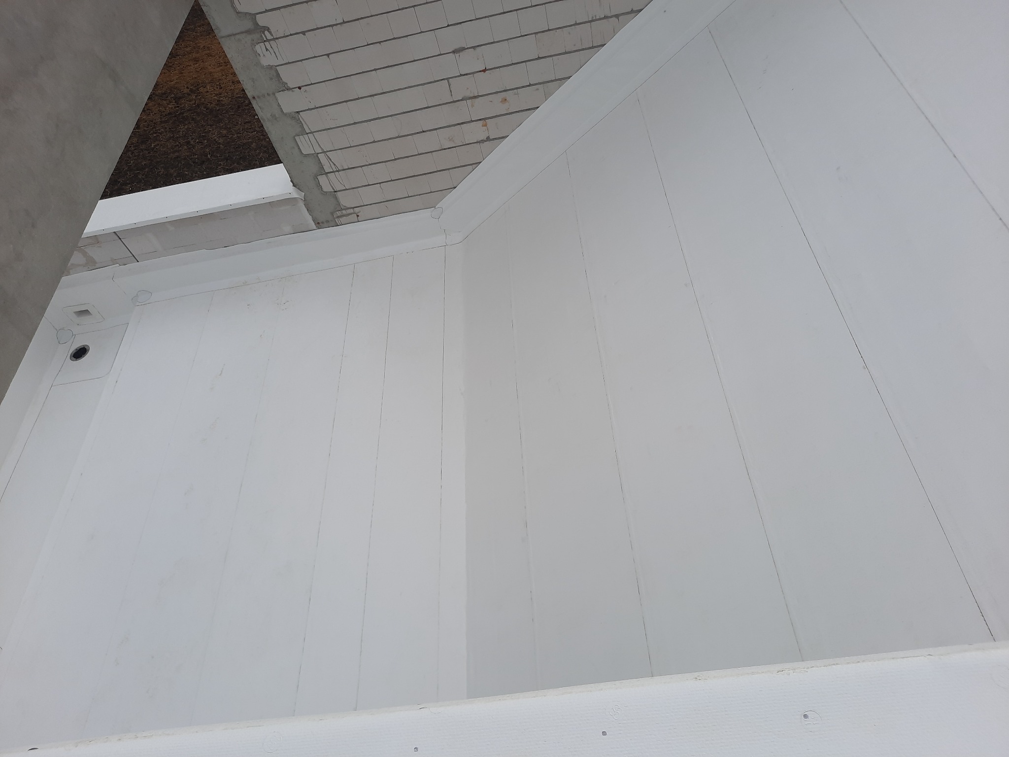 Widok z dołu na białą podbitkę dachową, z widocznymi elementami konstrukcji dachu i betonowym słupem w tle. Podbitka wykonana z paneli, z widocznymi łączeniami. Fragment surowego stropu z cegieł...
