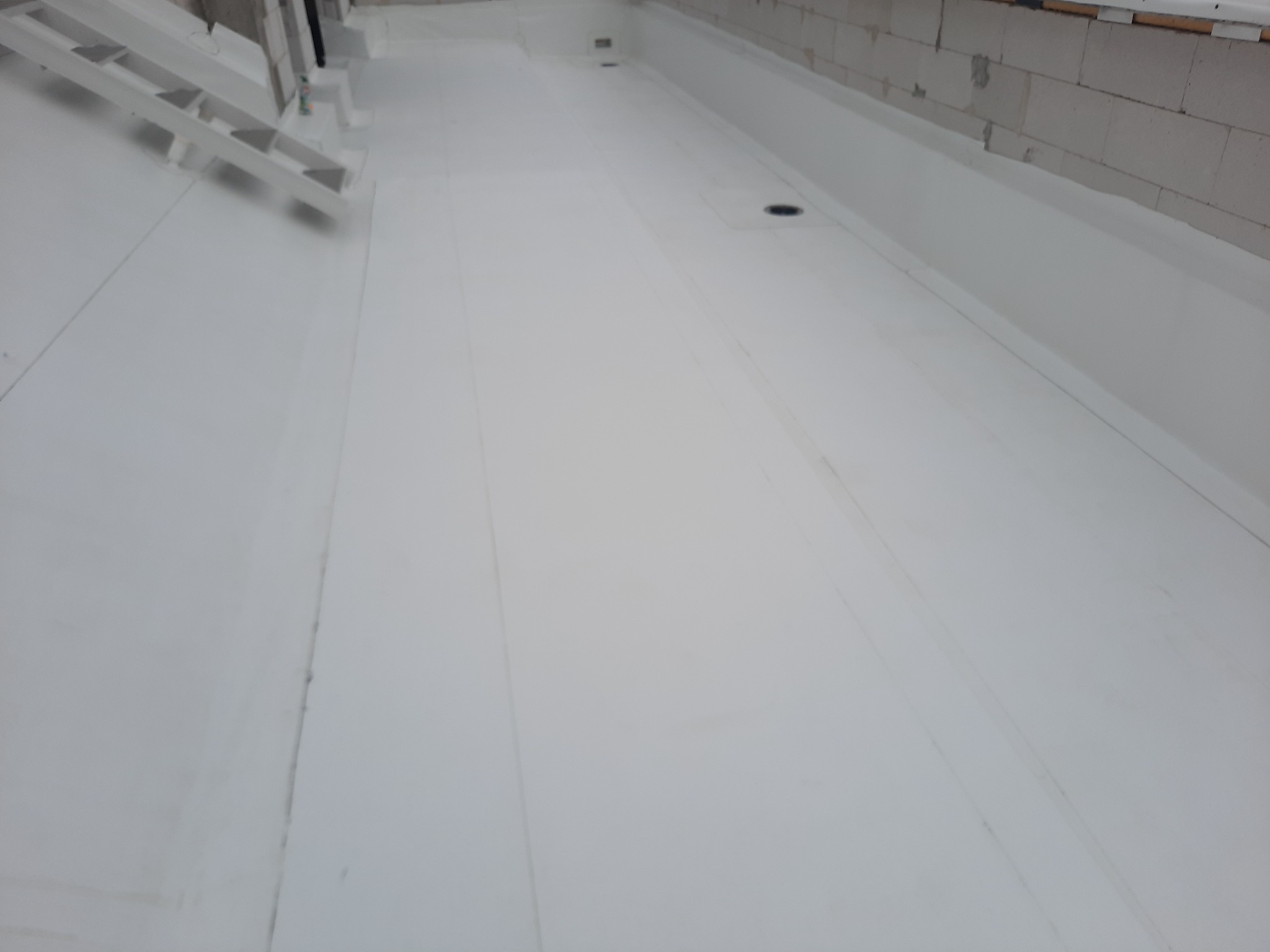 Biała, płaska powierzchnia dachu pokryta membraną hydroizolacyjną, z widocznymi wpustami dachowymi i fragmentem ściany z bloczków betonowych oraz białą drabiną w rogu.