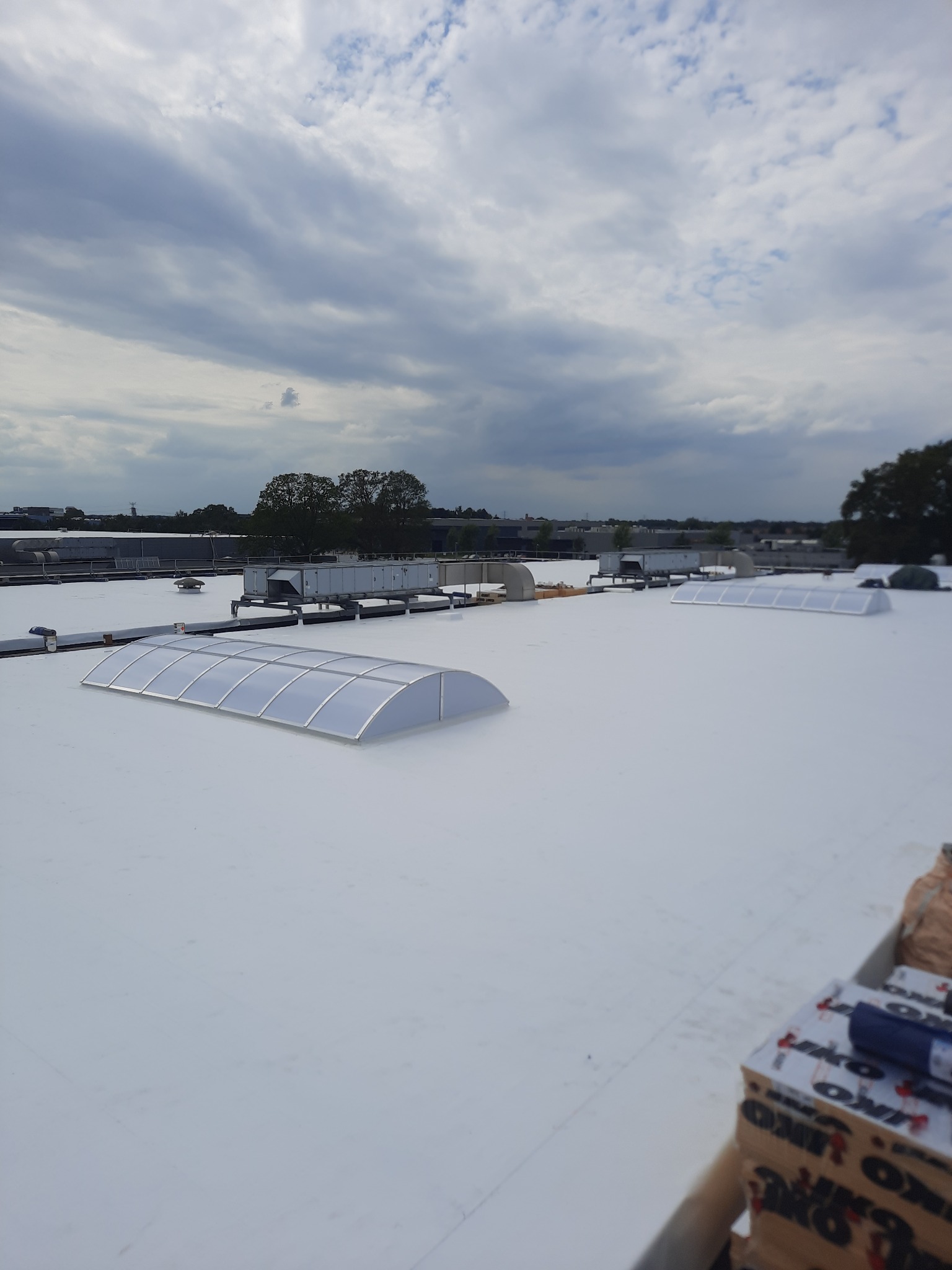 Biały dach płaski z świetlikami kopułowymi, widoczne urządzenia wentylacyjne i materiały izolacyjne w rogu, pochmurne niebo w tle.