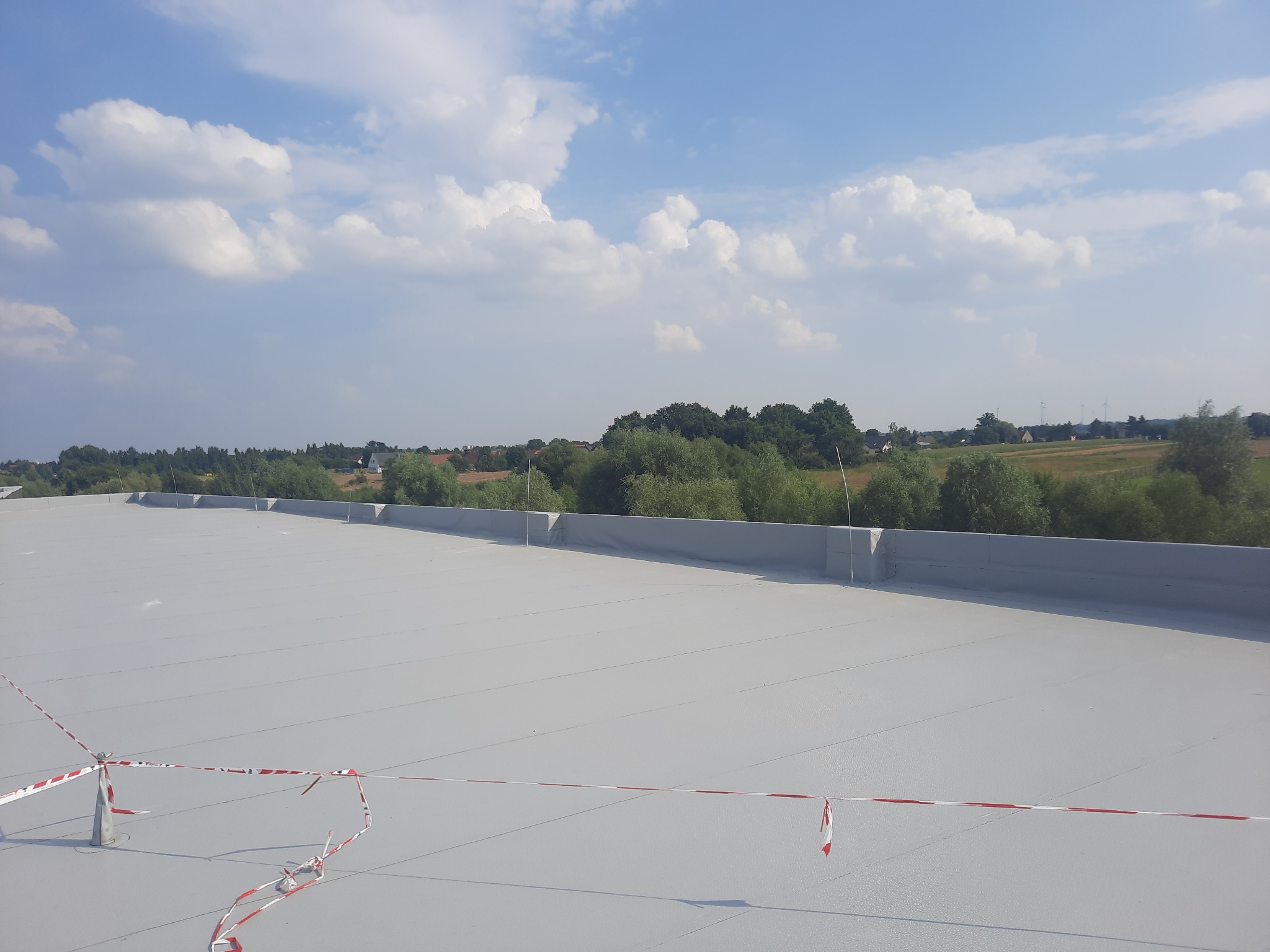 Szary, płaski dach przemysłowy z widocznym zabezpieczeniem w postaci taśmy ostrzegawczej, na tle zieleni i błękitnego nieba z chmurami.