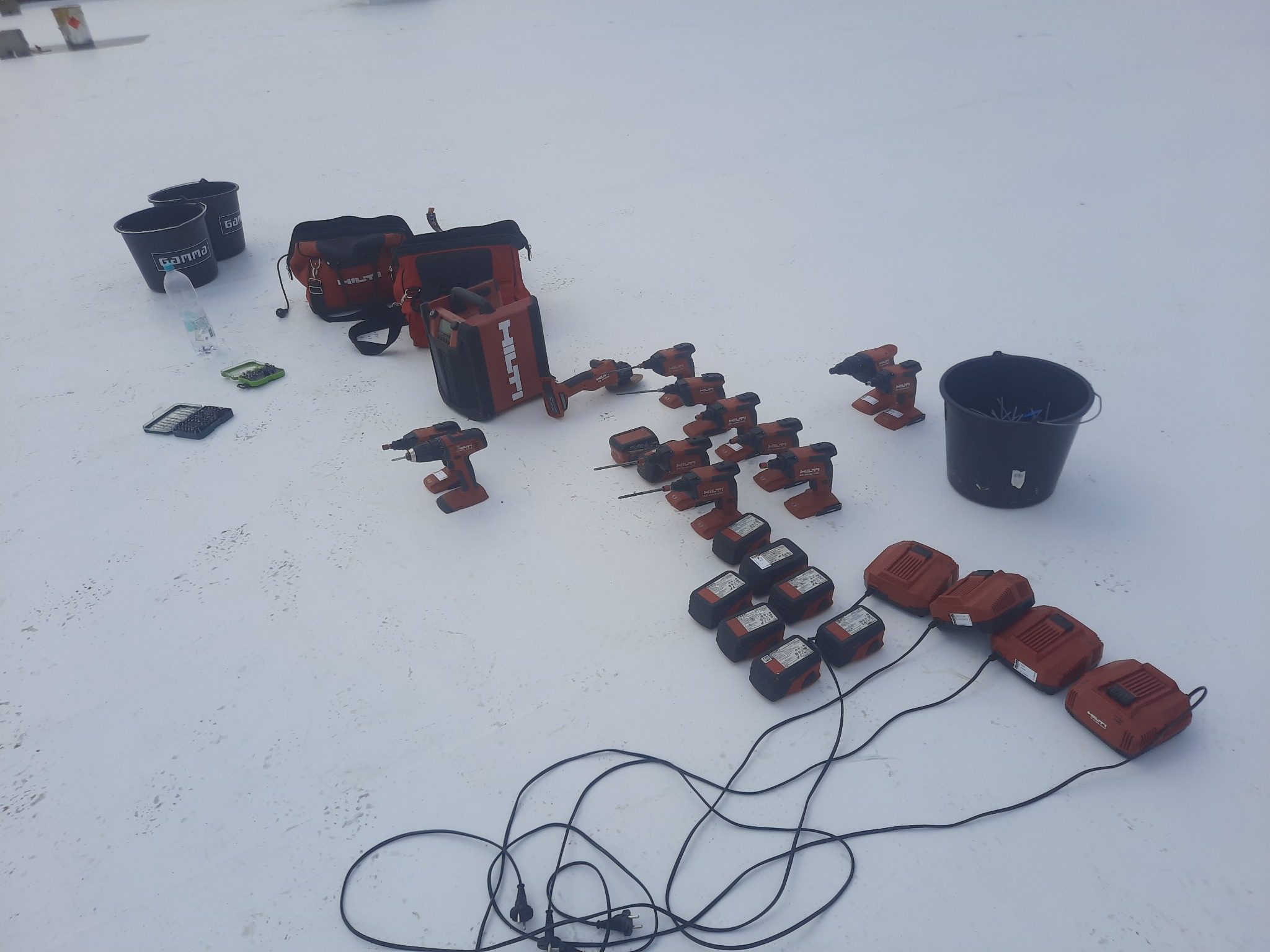 Rozłożone narzędzia dekarskie Hilti na białym dachu: wiertarki, akumulatory z ładowarkami, torby narzędziowe, dwa wiadra Gamma, butelka wody i małe pudełka z bitami.