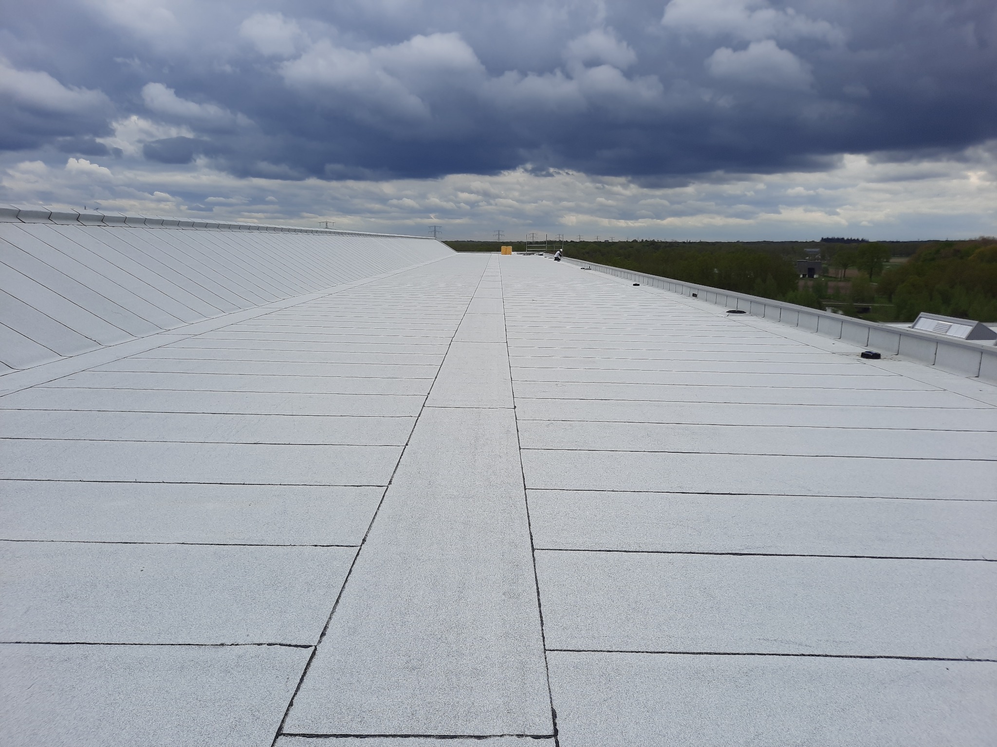 Długi, płaski dach pokryty szarymi panelami bitumicznymi, widok z góry, na horyzoncie linia drzew i pochmurne niebo.