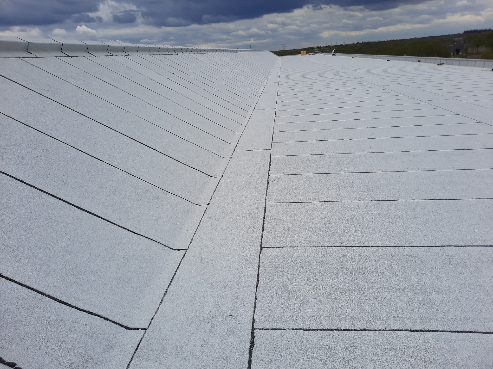 Widok z góry na płaski, pokryty jasną papą dach przemysłowy z widocznymi liniami łączeń, pochmurne niebo w tle.