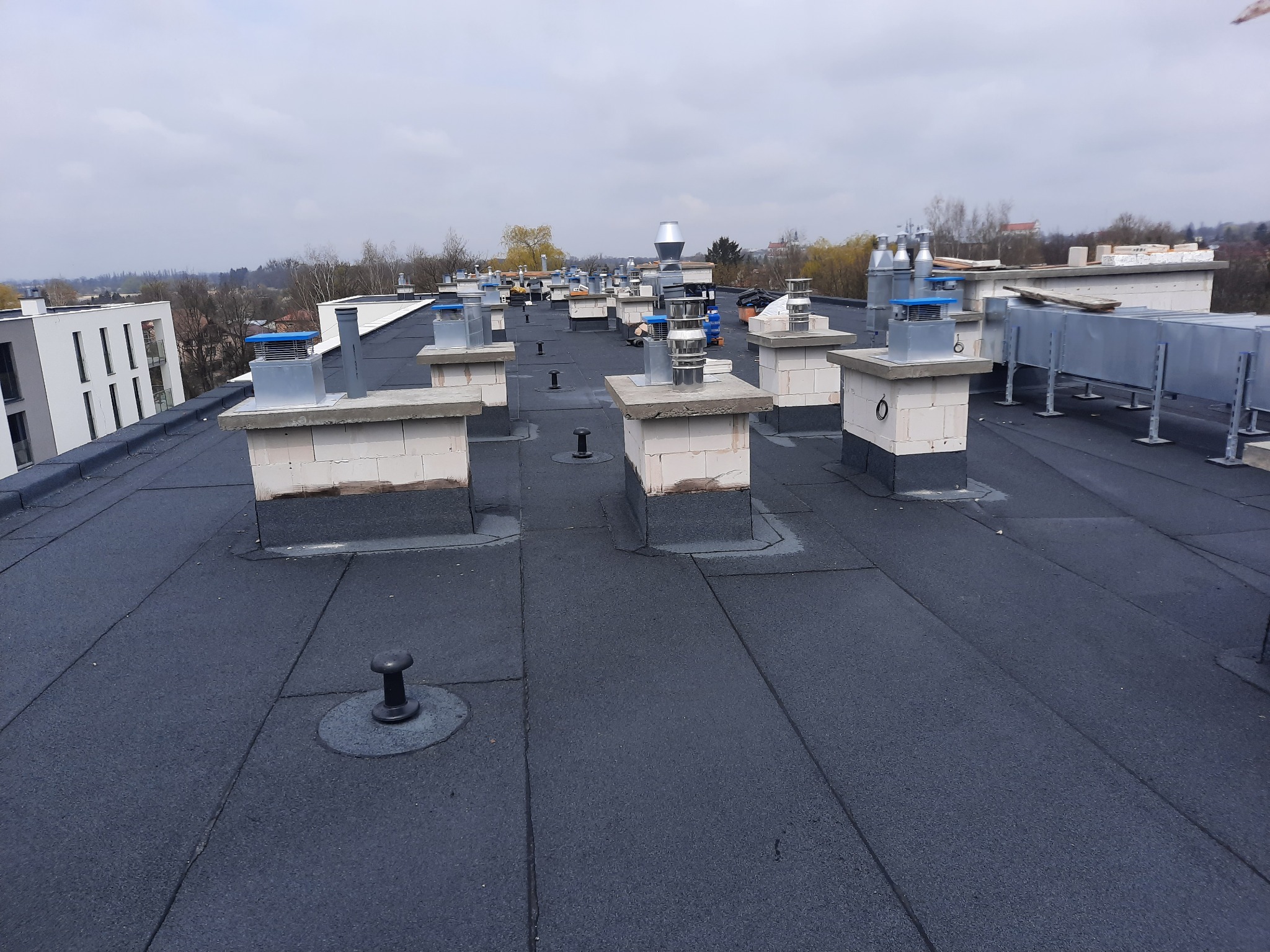 Widok z góry na płaski dach pokryty czarną papą z licznymi kominami wentylacyjnymi i odpowietrznikami, w tle zabudowa miejska i pochmurne niebo.