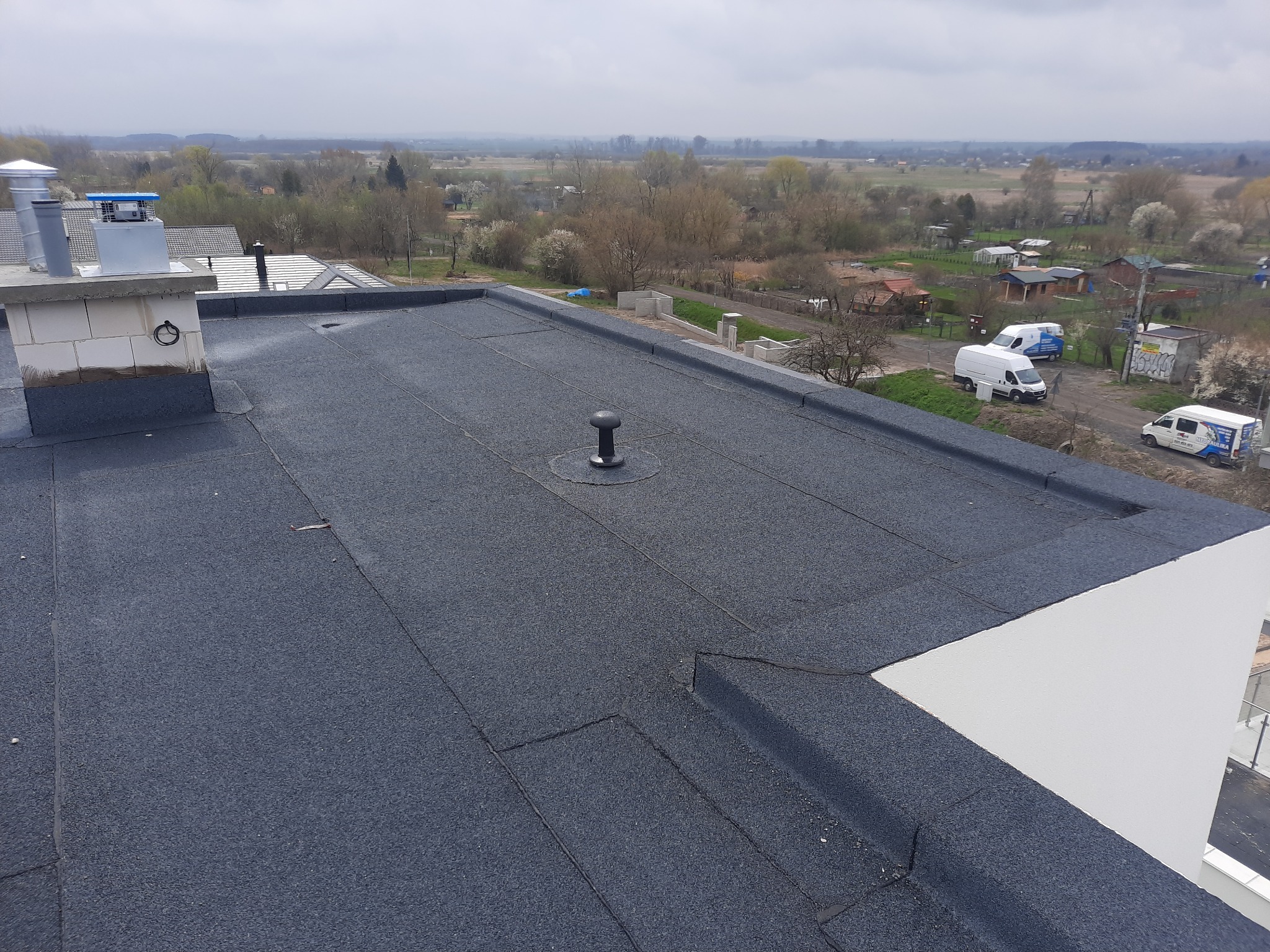 Widok z góry na płaski dach pokryty papą termozgrzewalną, z kominem z cegły i elementami wentylacyjnymi, w tle zabudowa jednorodzinna i wiosenny krajobraz.