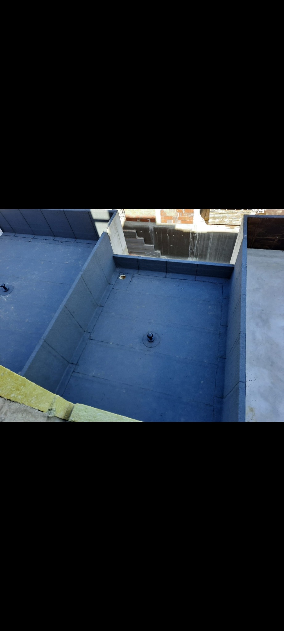 Płaski dach pokryty szarą papą z widocznymi detalami mocowania i obróbkami blacharskimi, widok z góry na fragment dachu z otoczeniem i budynkiem w tle.