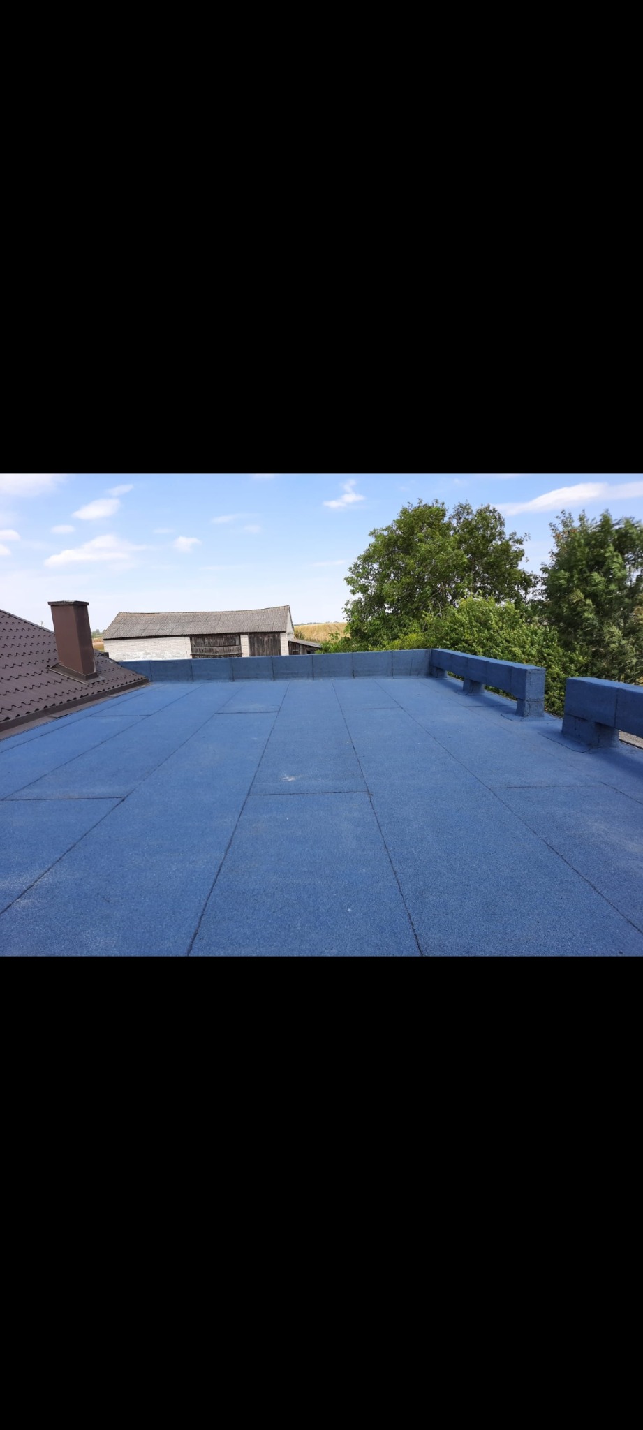 Niebieski dach pokryty płytami z widocznym kominem budynku obok i budynkiem gospodarczym w tle.