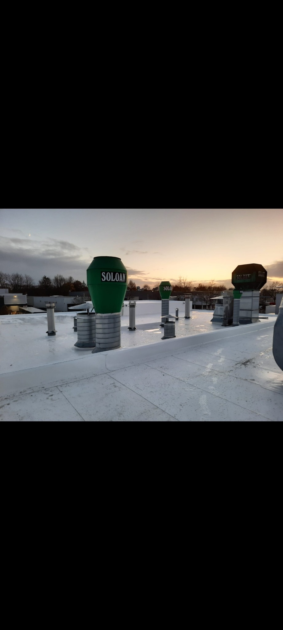 Biały, płaski dach komercyjny z kilkoma zielonymi kominami wentylacyjnymi z napisem 'SOLOAN' i metalowymi podstawami, sfotografowany o zmierzchu.