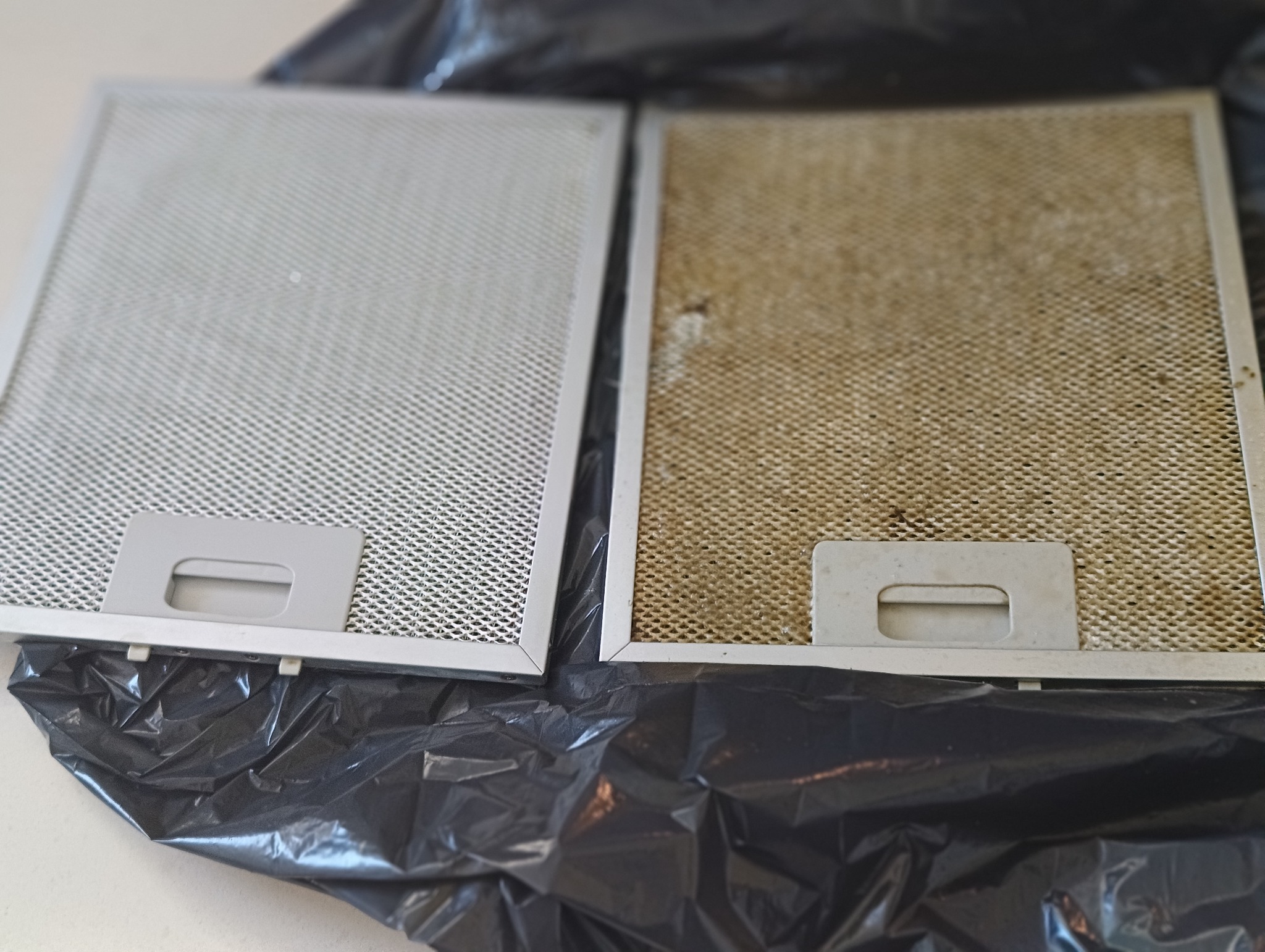 Filtr okapu kuchennego przed i po czyszczeniu, ukazujący różnicę w zabrudzeniu. Jeden filtr jest czysty, drugi pokryty tłustym brudem, leżą na czarnej folii.