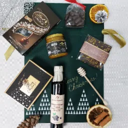 Świąteczny kosz prezentowy z czekoladkami Lindt, miodem kremowanym z cytryną i imbirem, syropem porzeczkowym i dekoracjami na zielonej serwetce z choinkami.