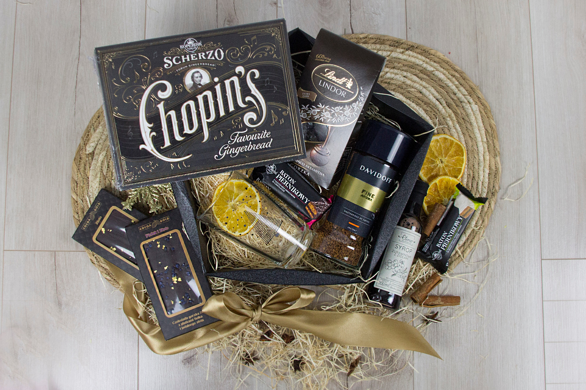 Elegancki, czarny kosz prezentowy wypełniony słodyczami, kawą Davidoff i syropem, ozdobiony złotą wstążką i suszonymi pomarańczami, ułożony na plecionym podkładzie.