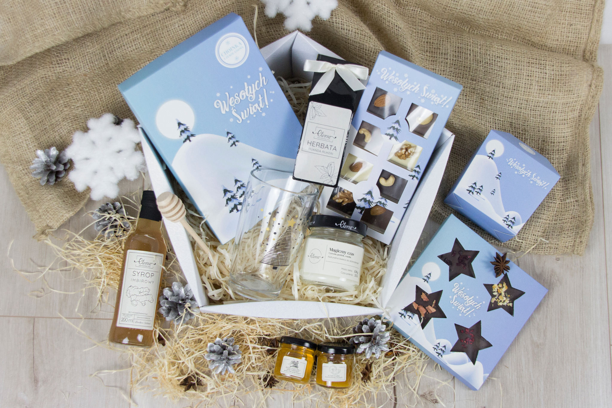 Świąteczny zestaw upominkowy w białym pudełku, zawierający syrop imbirowy, herbatę, świecę, miód, szklankę z choinką i czekoladki w pudełkach z motywem zimowym.