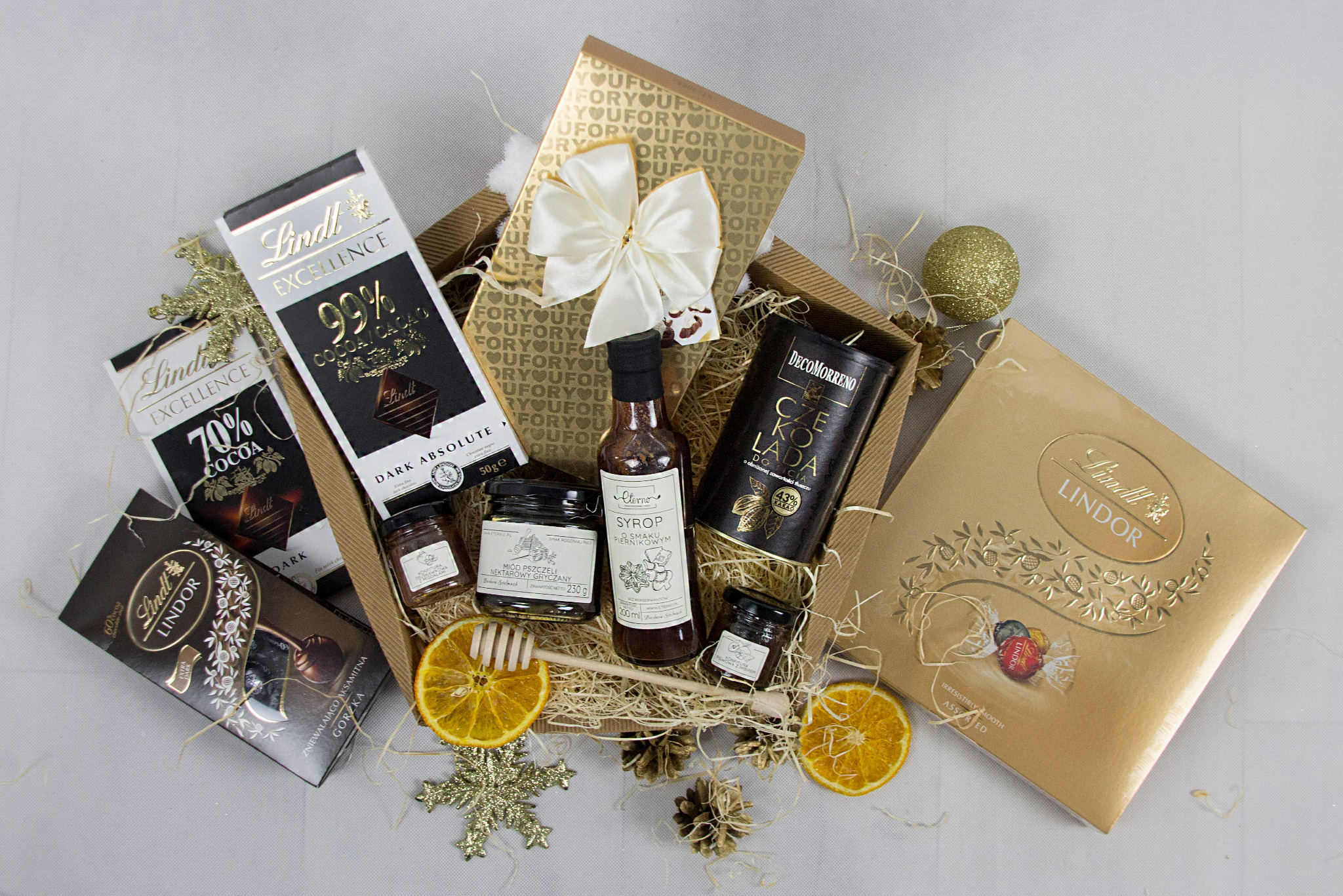 Zestaw prezentowy z czekoladami Lindt, miodem, syropem piernikowym, plastrami pomarańczy i złotą ozdobą w kształcie płatka śniegu, umieszczony w brązowym koszu wypełnionym wiórkami.