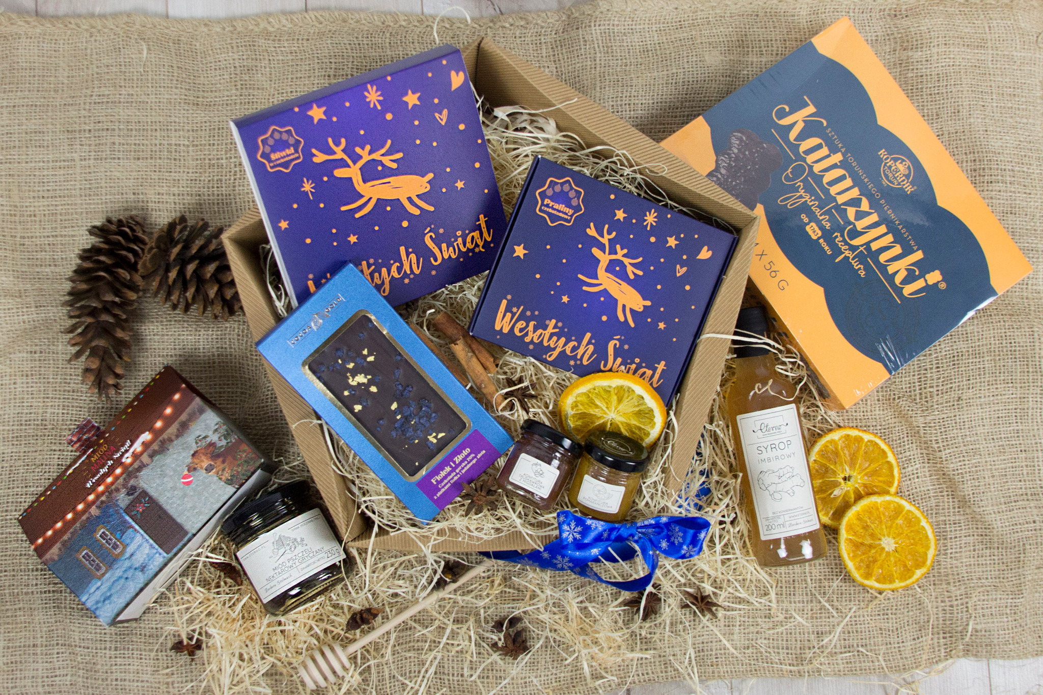 Świąteczny zestaw upominkowy z piernikami, czekoladą, miodem i syropem imbirowym, zapakowany w kartonowe pudełko z dekoracjami, na jutowym tle.
