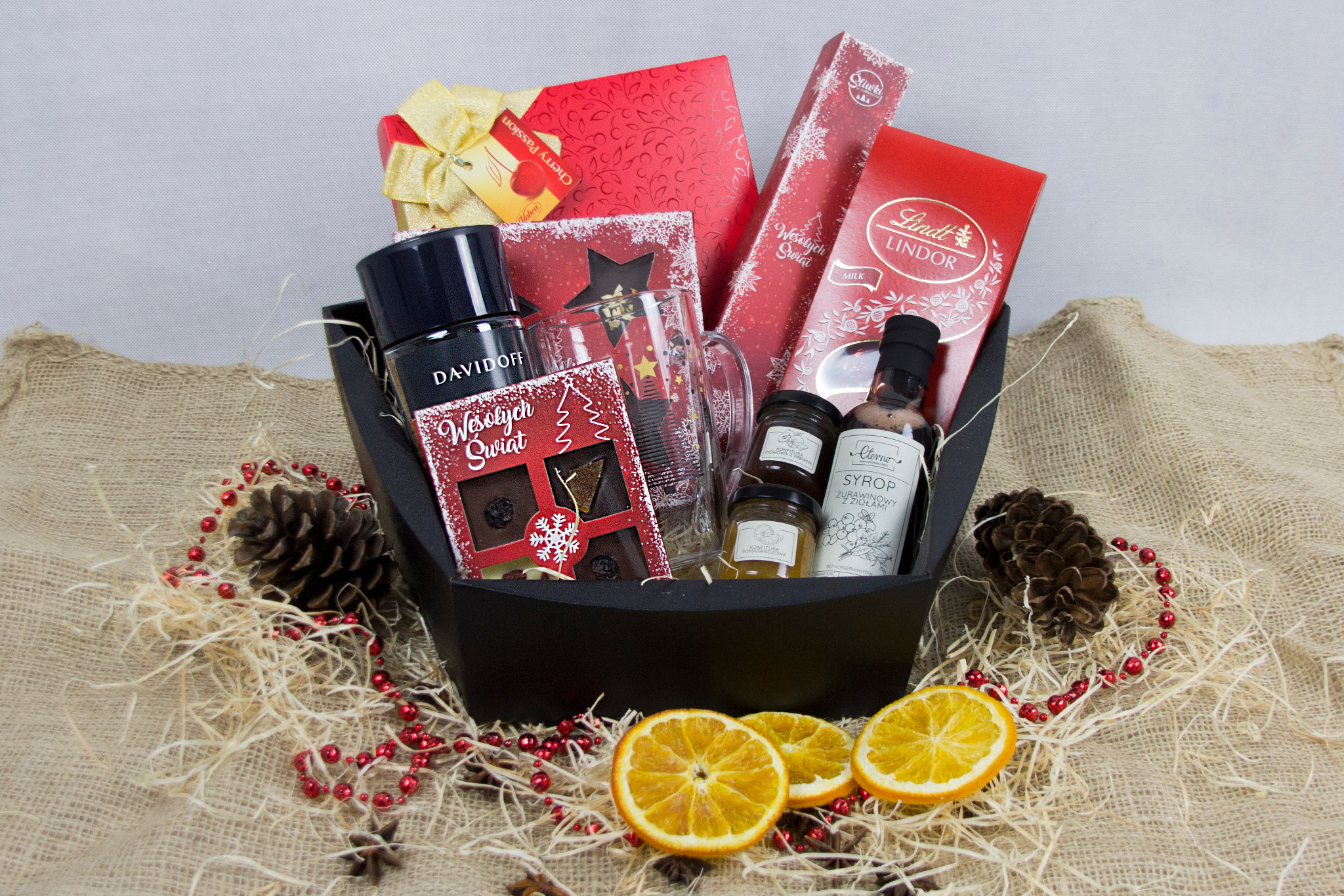 Czarny kosz prezentowy wypełniony słodyczami, perfumami, syropem, miodem i kubkiem świątecznym, ozdobiony plasterkami pomarańczy, szyszkami i czerwonymi koralikami na tle jutowym.