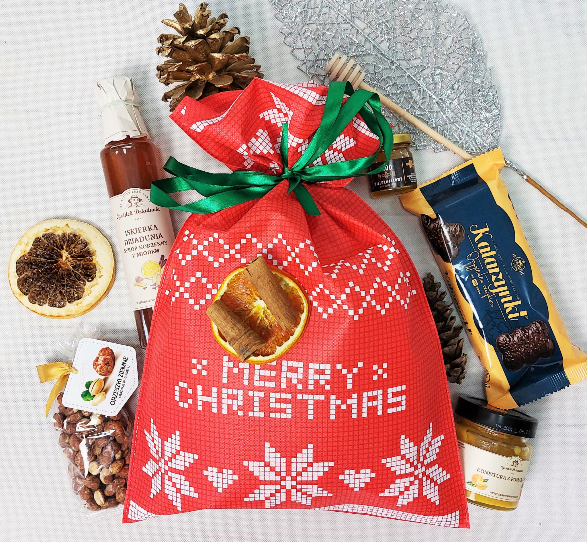Czerwony worek prezentowy z napisem Merry Christmas i dekoracją z suszonej pomarańczy z cynamonem, otoczony miodami, konfiturą, piernikami Katarzynki i orzeszkami w karmelu na białym tle.