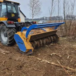 Żółto-czarny traktor JCB z niebiesko-żółtą frezarką do pni Bomford na polu z pozostałościami po wycince drzew, w tle budynek i drzewa.