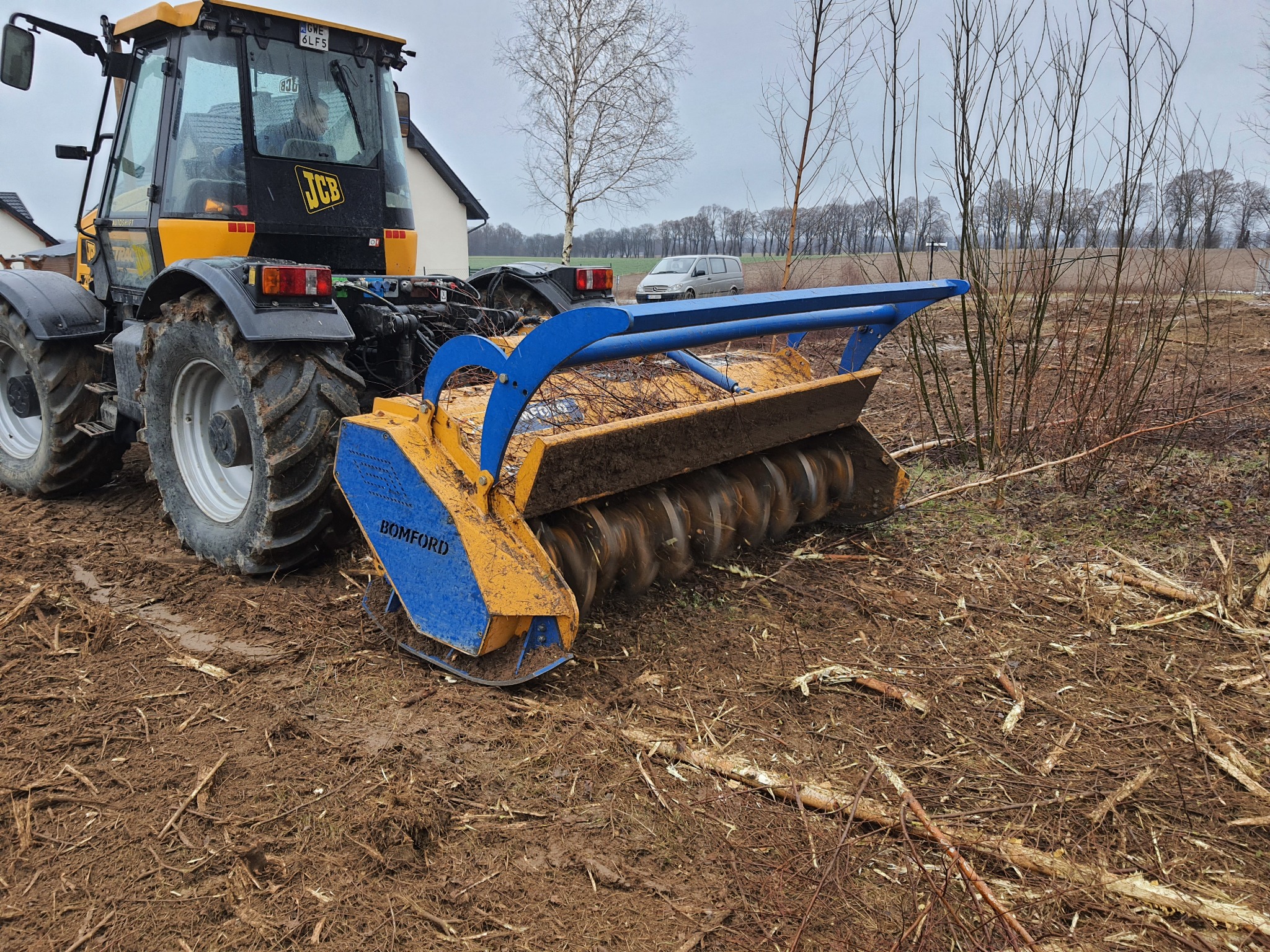 Żółto-czarny traktor JCB z niebiesko-żółtą frezarką do pni Bomford na polu z pozostałościami po wycince drzew, w tle budynek i drzewa.