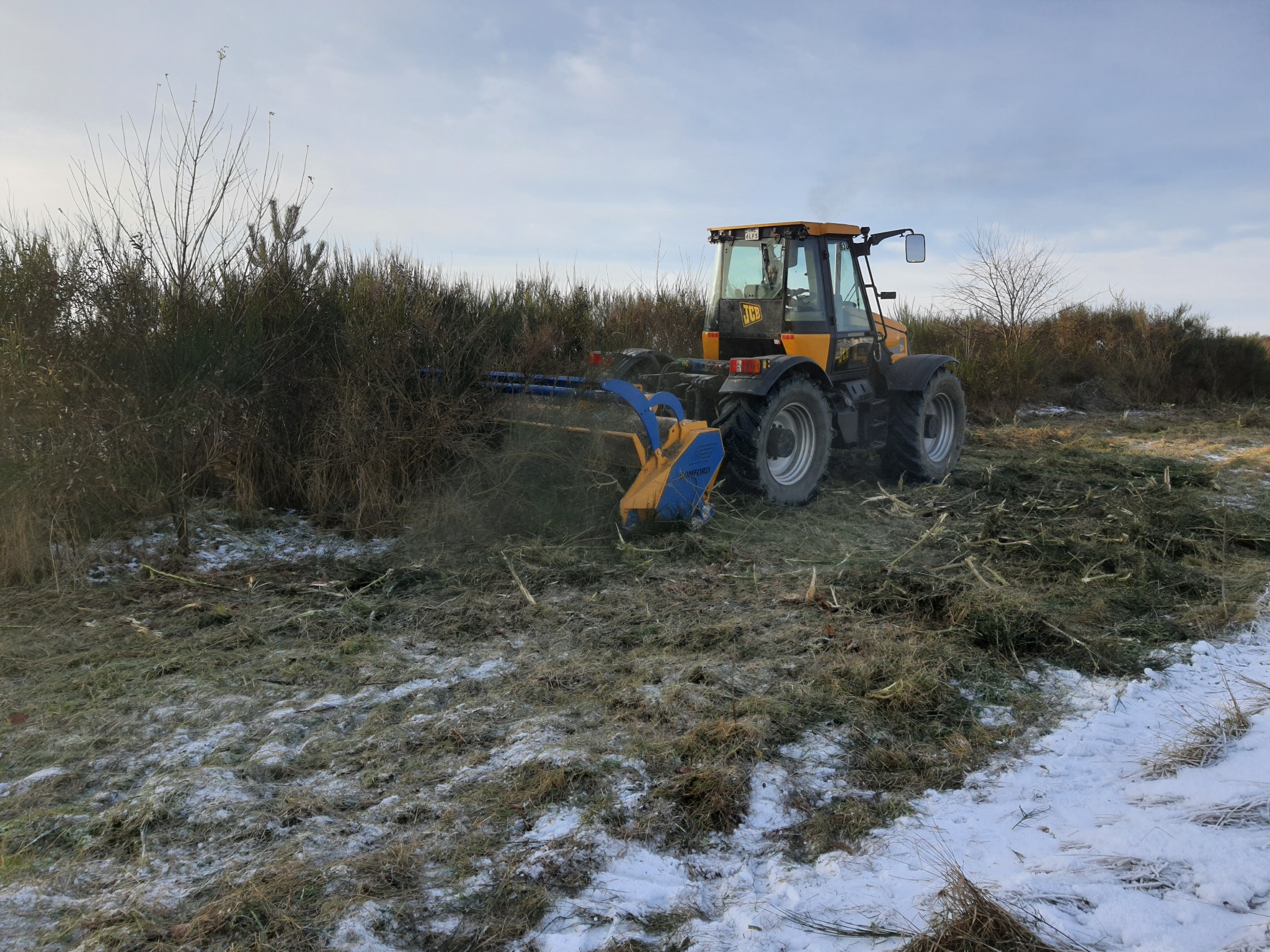 Żółty traktor z kosiarką rotacyjną tnie krzewy na polu, w tle pochmurne niebo.