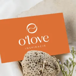 Pomarańczowa wizytówka z białym logo i napisem 'o'love inspiracje', oparta o porowaty kamień, z delikatnymi kwiatami na tle białej satyny.