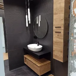 Nowoczesna łazienka z czarnymi płytkami, owalną białą umywalką nablatową, szafką z drewnopodobnym fornirem i okrągłym lustrem; widoczny surowy fragment ściany przed wykończeniem.