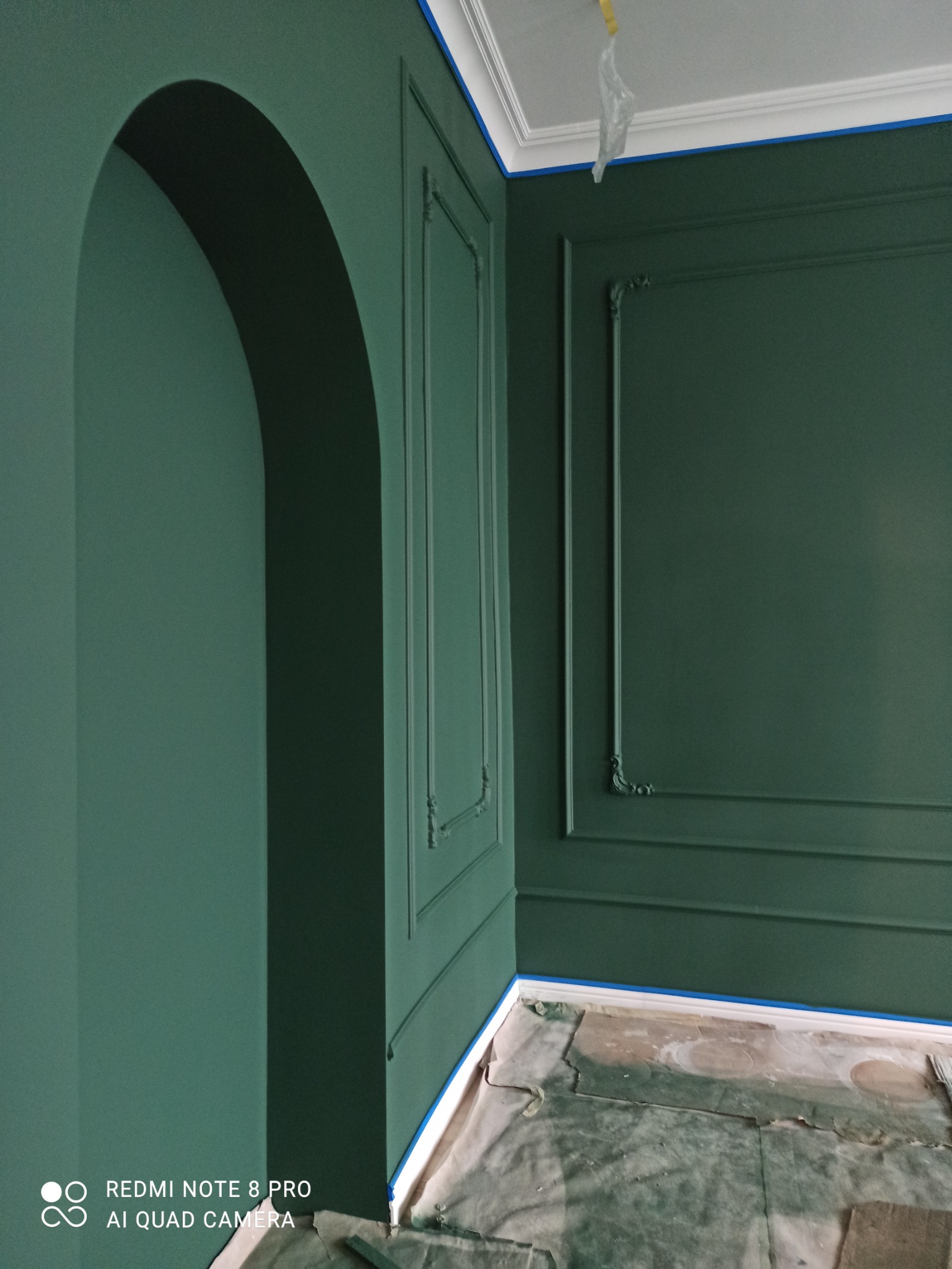 Ściana pomalowana na ciemnozielony kolor z ozdobnymi listwami, widoczne zabezpieczenia taśmą malarską przy suficie i podłodze, oraz zakryta podłoga folią ochronną.