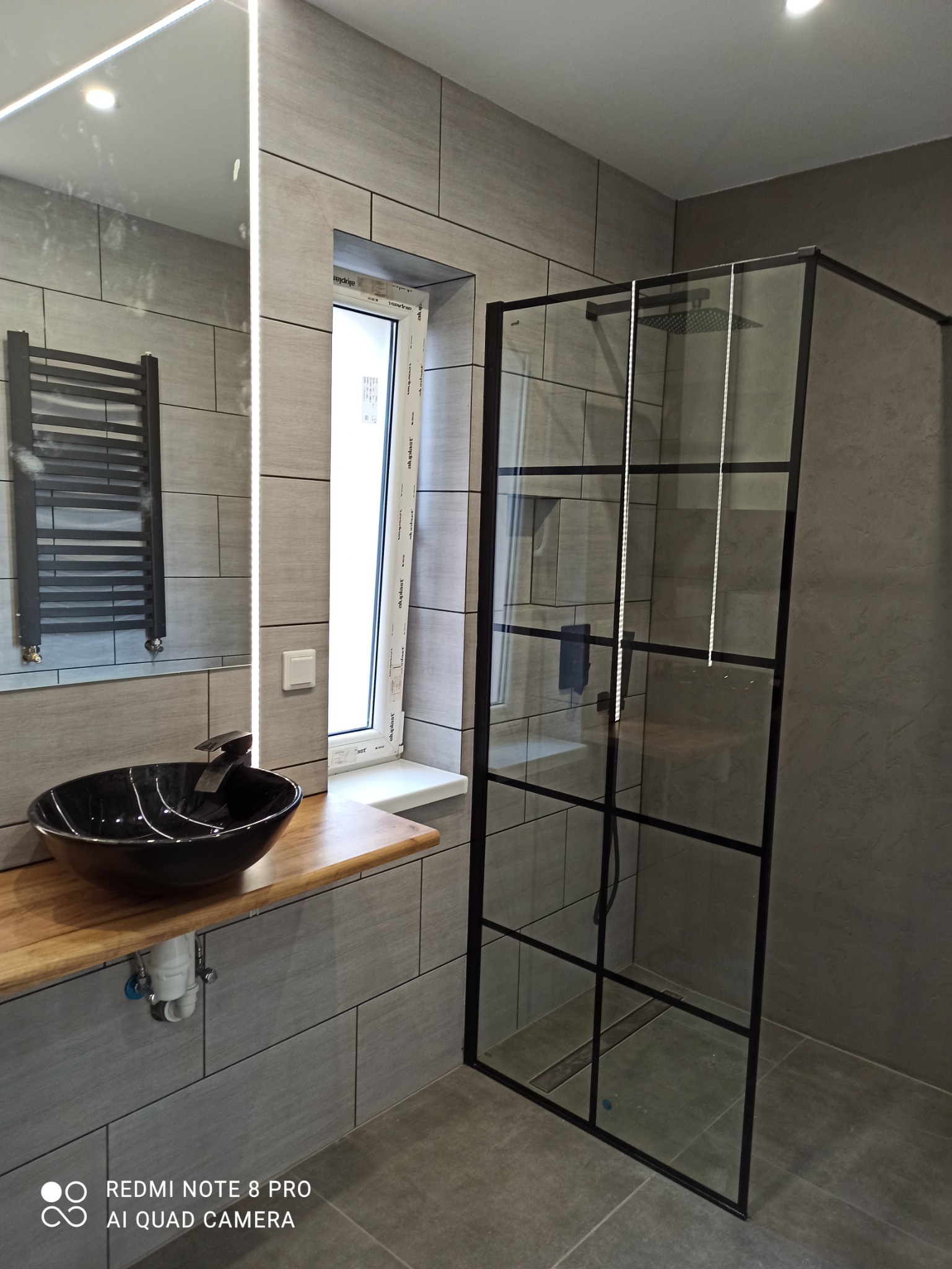 Nowoczesna łazienka z szarymi płytkami, czarną umywalką na drewnianym blacie, grzejnikiem drabinkowym w lustrze i kabiną prysznicową z czarnymi ramami.