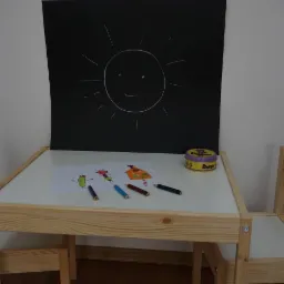 Dziecięcy stolik z rysunkiem domu i postaci, kolorowe kredki, tablica z narysowanym słońcem i puszka kredek w tle.