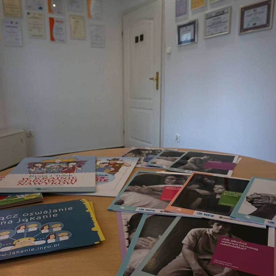 Stół z materiałami edukacyjnymi, książeczkami i ulotkami o tematyce logopedycznej, w tle ściana z dyplomami i certyfikatami oraz białe drzwi.