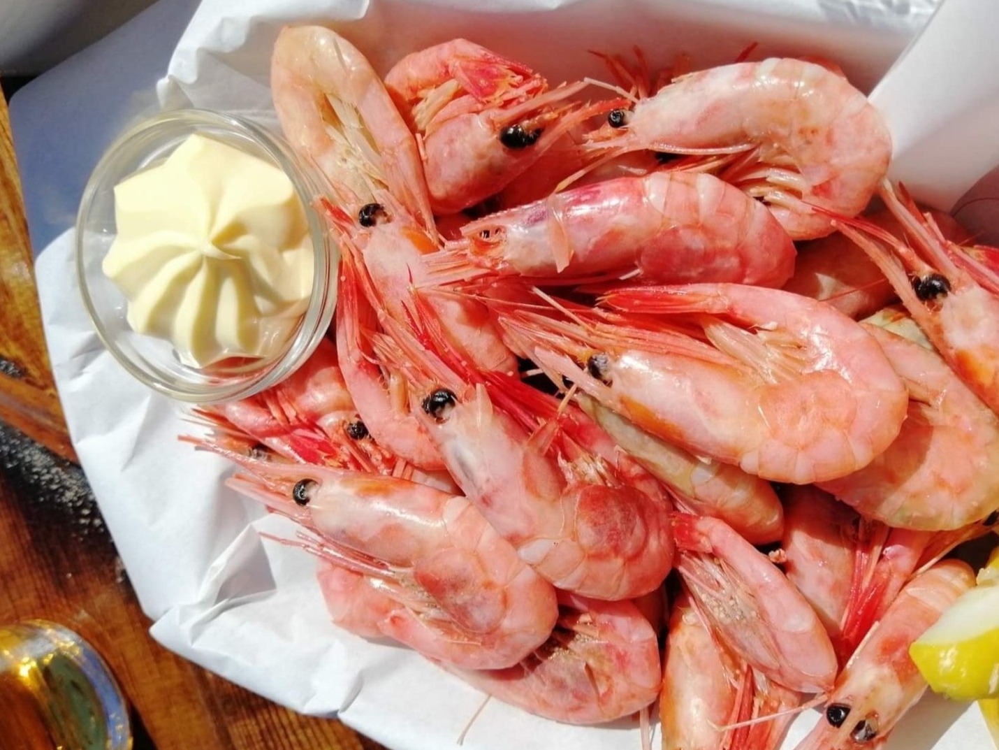 Ułożone na papierze różowe krewetki z czerwonymi czułkami, obok miseczka z kremowym sosem i kawałek cytryny, widoczne na drewnianym stole.