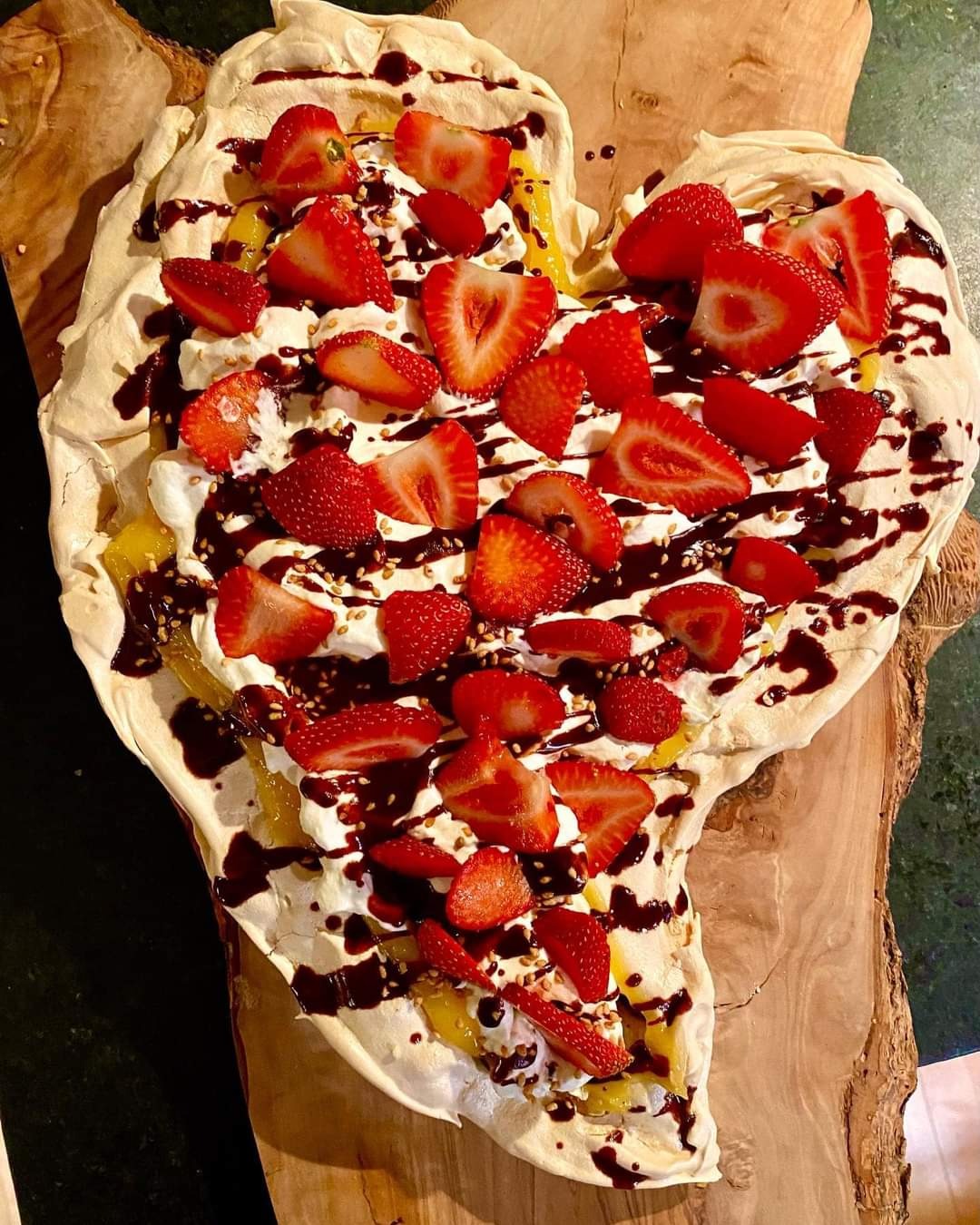 Pavlova w kształcie serca na drewnianej desce, udekorowana bitą śmietaną, świeżymi truskawkami, sosem czekoladowym i posypką z sezamu.