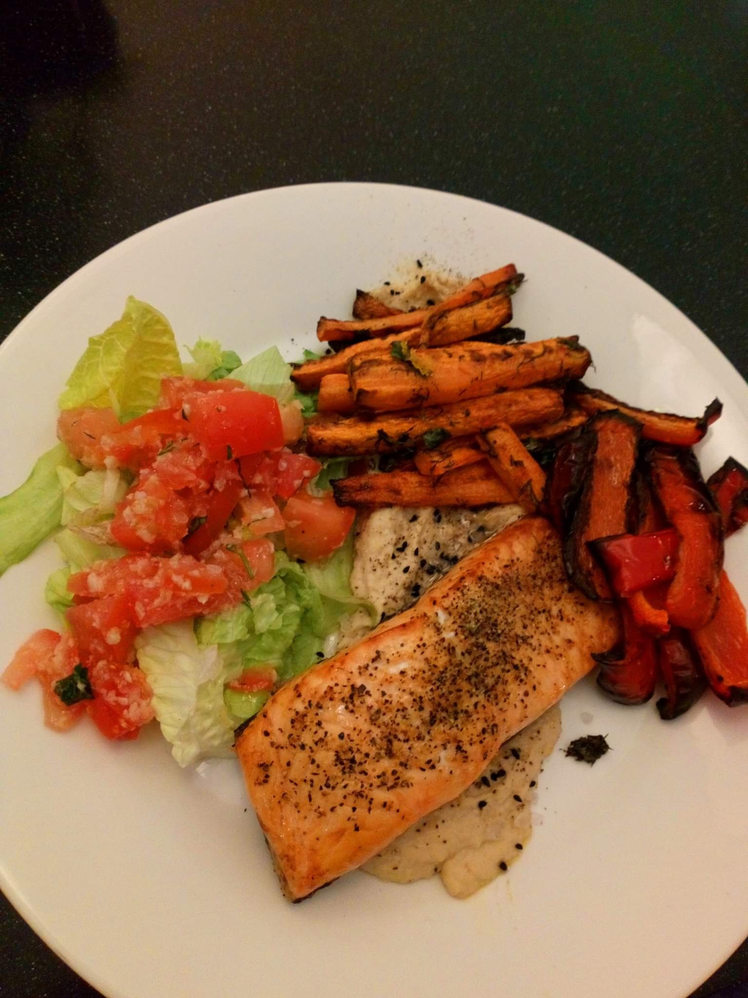 Porcja grillowanego łososia z posypką z czarnego pieprzu, podana na białym talerzu z puree, sałatką z pomidorów i sałaty oraz pieczonymi frytkami z batatów i kawałkami papryki.