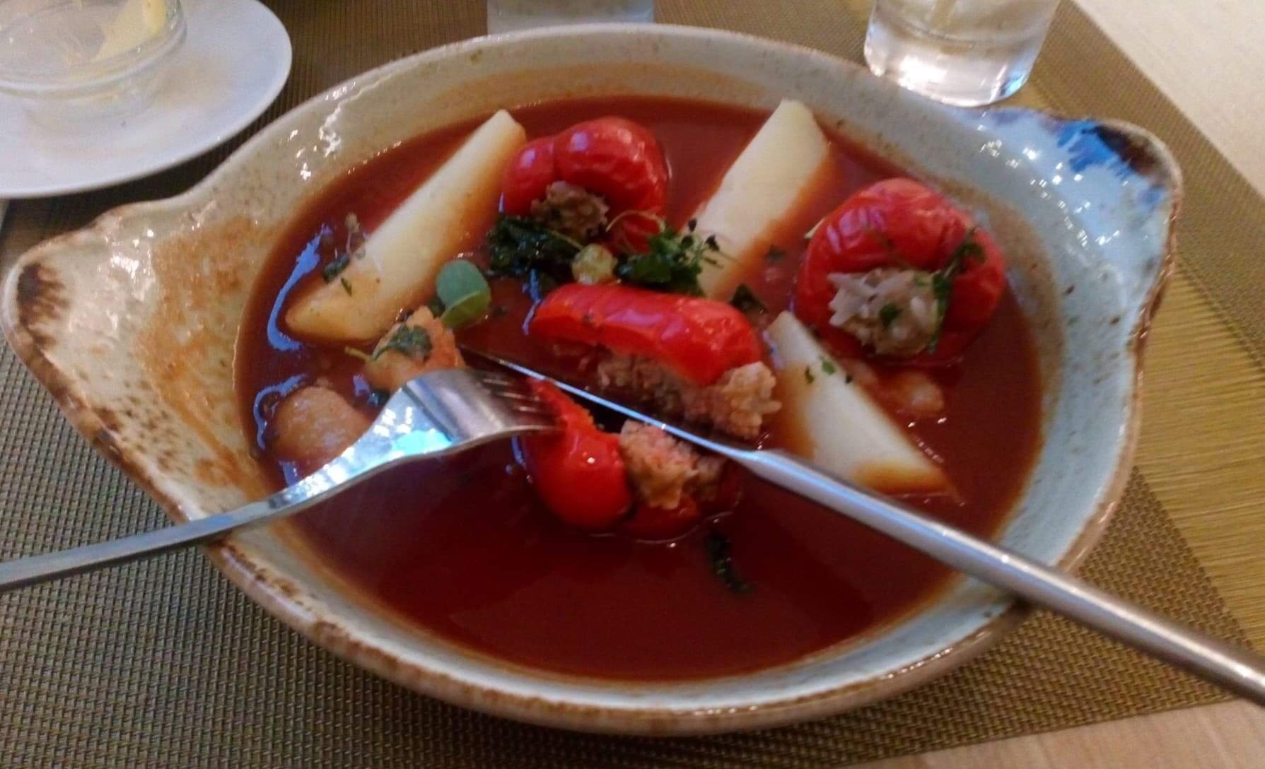 Głębia talerza z zupą pomidorową, w której pływają faszerowane papryki, ziemniaki i zioła, częściowo spożywana przy użyciu widelca i noża.