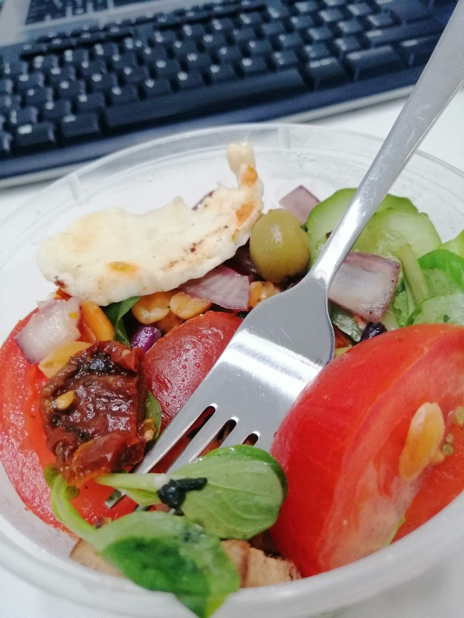 Sałatka w plastikowym pojemniku na biurku z widoczną klawiaturą, zawiera pomidory, ogórki, oliwki, czerwoną cebulę i kawałek pieczywa, w sałatce wbity widelec.