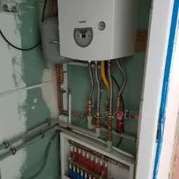 Biały piec gazowy Termet zamontowany na ścianie w trakcie instalacji, widoczne liczne rury miedziane i elastyczne przewody, rozdzielacz z czerwono-niebieskimi pętlami ogrzewania podłogowego, surowe...