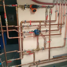 Rozbudowana instalacja hydrauliczna z rur miedzianych z zaworami Flowline i pompami obiegowymi LFP, zamocowana do ściany, widoczna butelka wody na podłodze.
