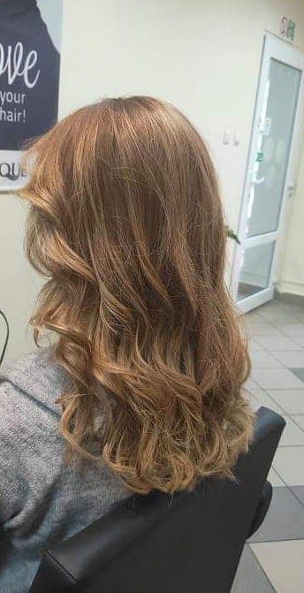 Kobieta z długimi, brązowymi włosami z delikatnymi falami, siedząca na czarnym krześle w salonie.