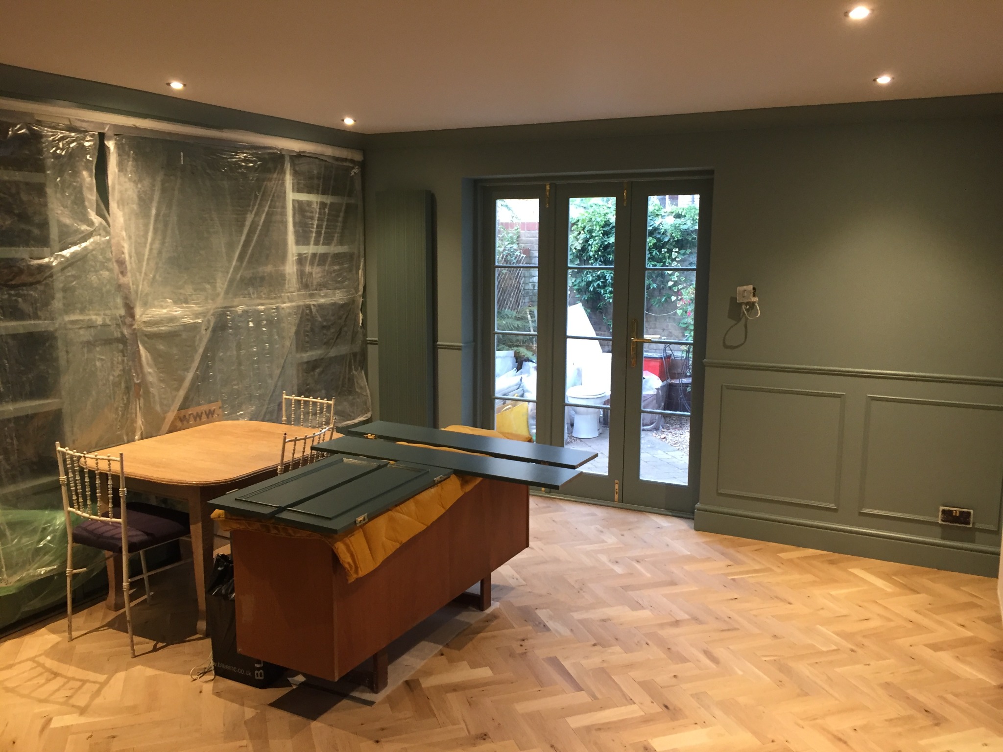 Pokój w trakcie prac: ściany pomalowane na oliwkowy kolor, podłoga z parkietu ułożonego w jodełkę, okno zabezpieczone taśmą malarską, na komodzie leżą pomalowane elementy stolarki.