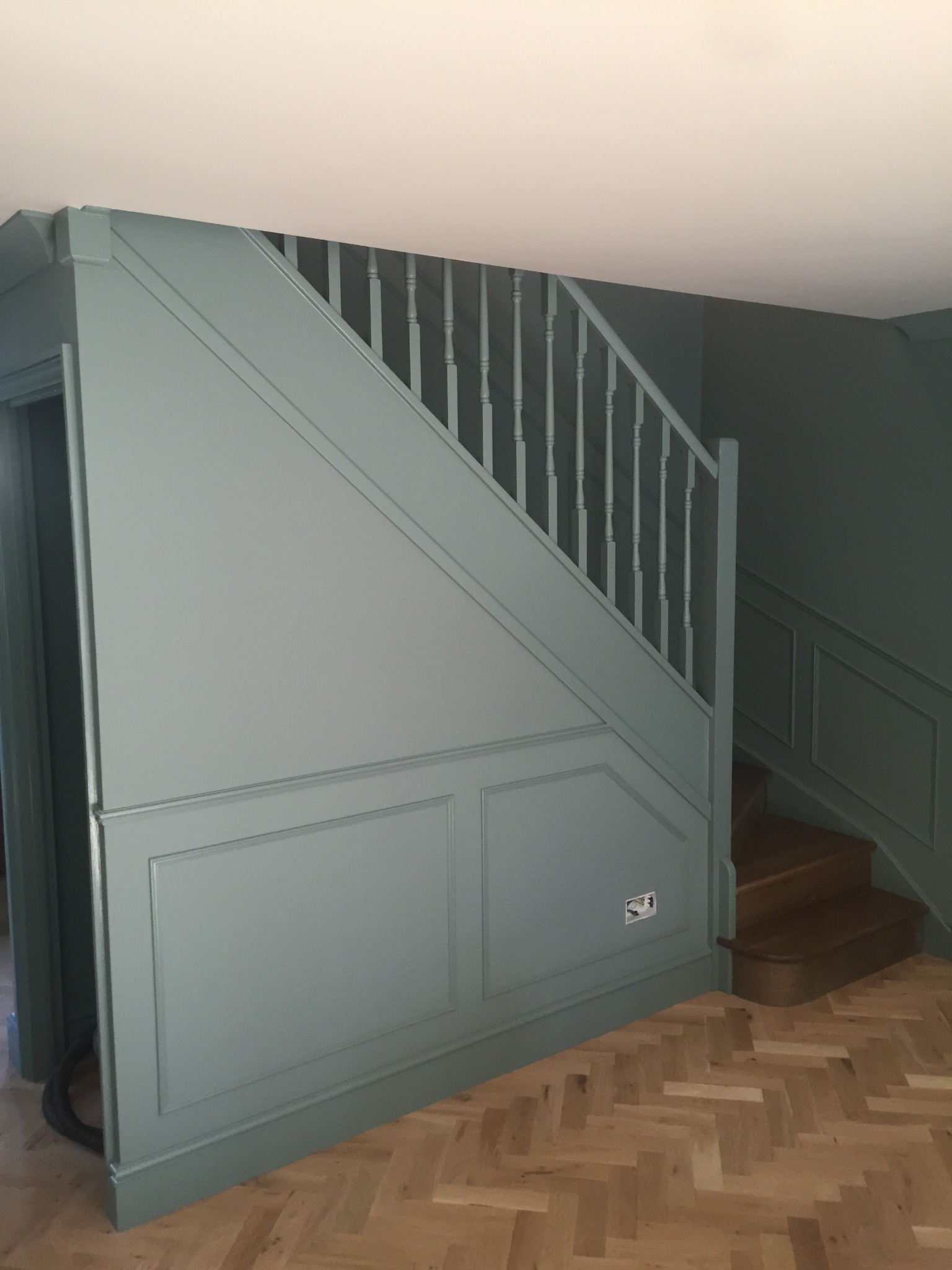 Klatka schodowa z drewnianymi schodami i ścianami w odcieniach zieleni z dekoracyjnymi panelami, parkiet ułożony w jodełkę.